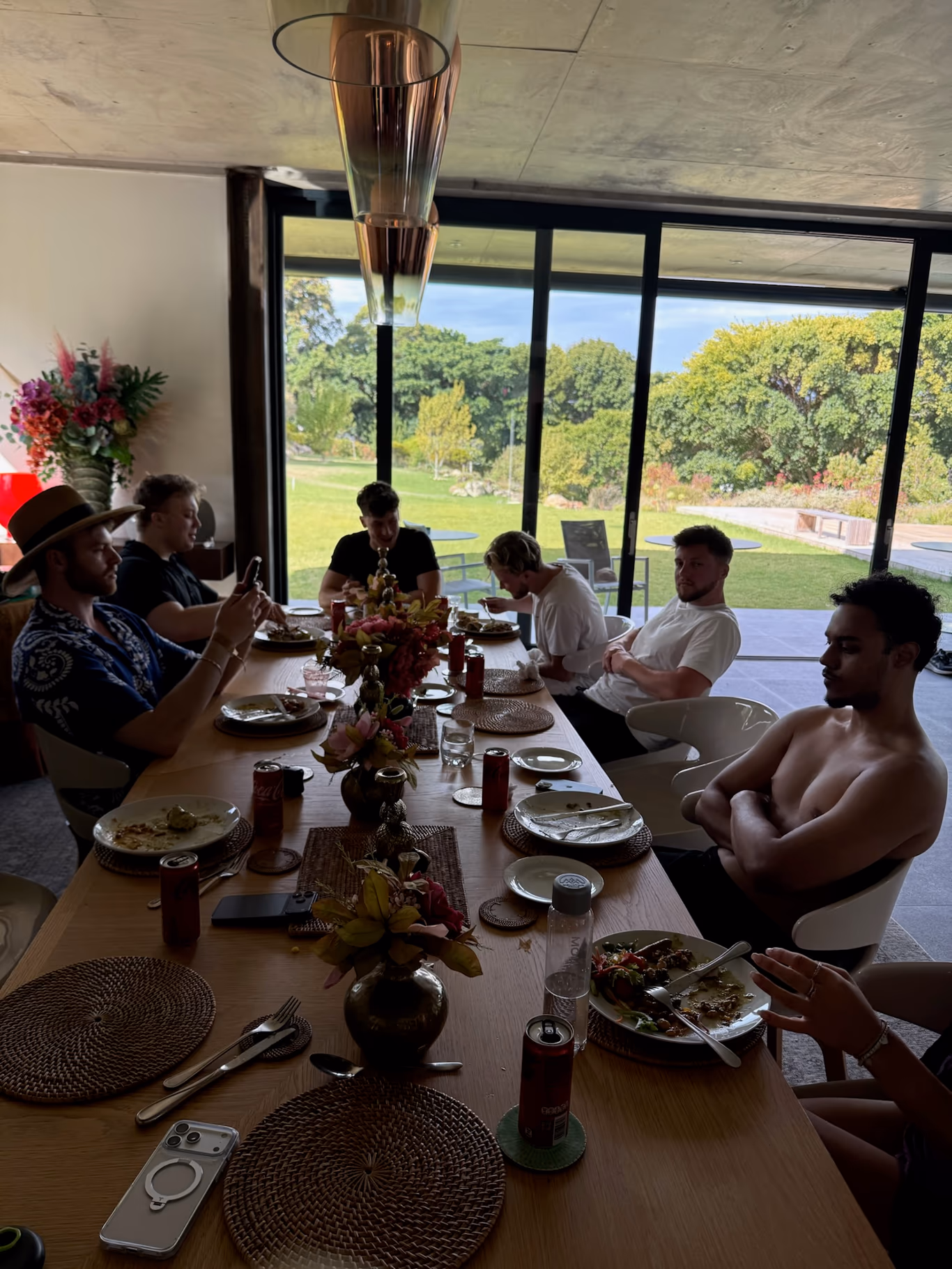Founders gathered around a long wooden table for a midday meal, floor-to-ceiling windows behind them framing a green garden
