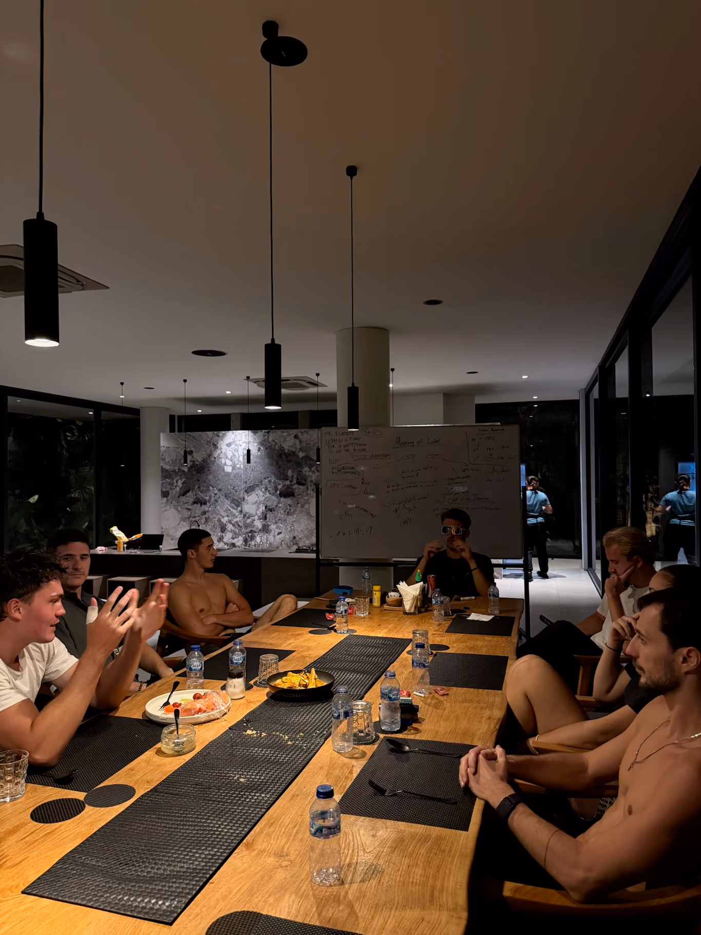 Founders seated around a long table late at night at a Helix villa, one mid-gesture, a whiteboard of notes behind them