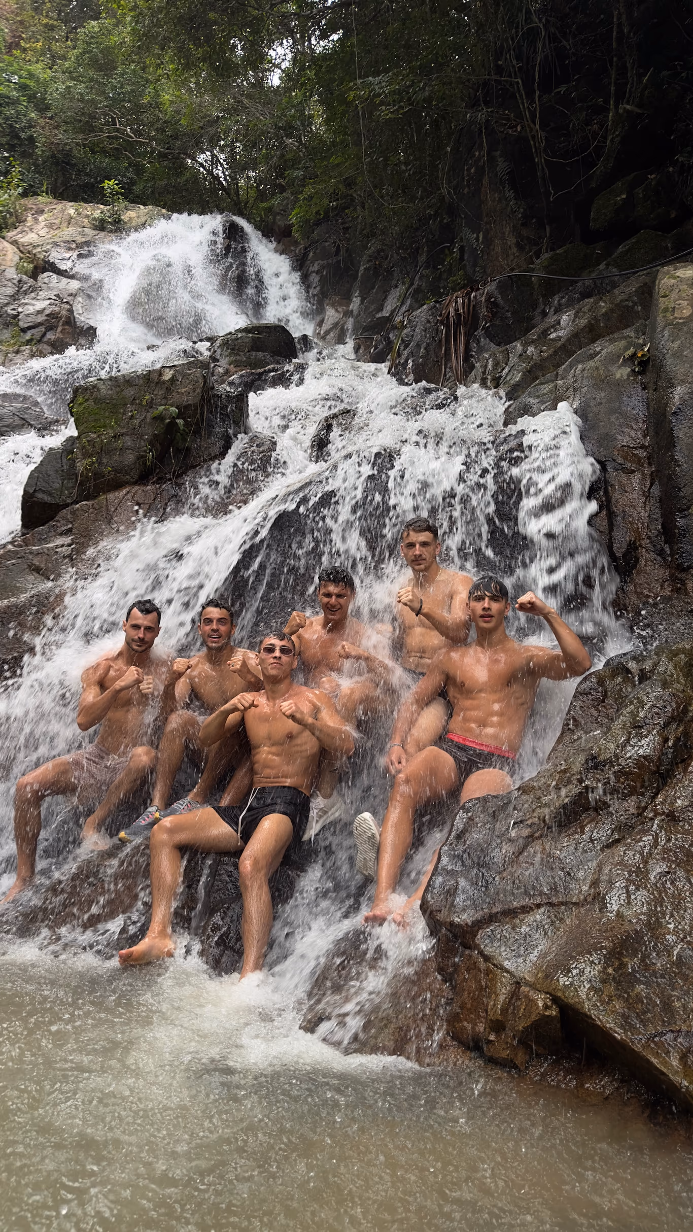 A founder travel group posing together in swim trunks under a cascading waterfall on a Helix trip — off the itinerary
